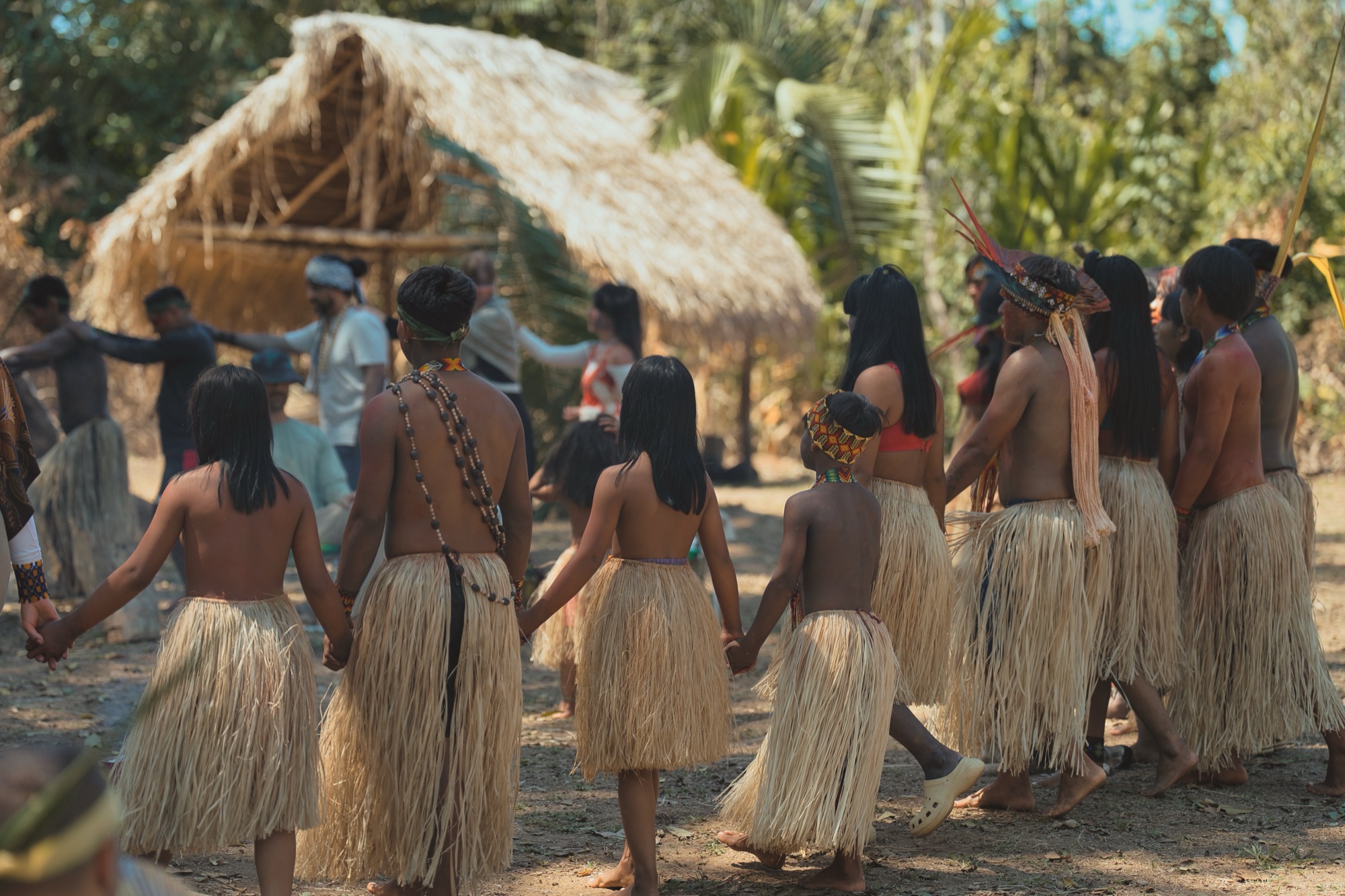 The community walks hand in hand toward the palm-thatched maloca